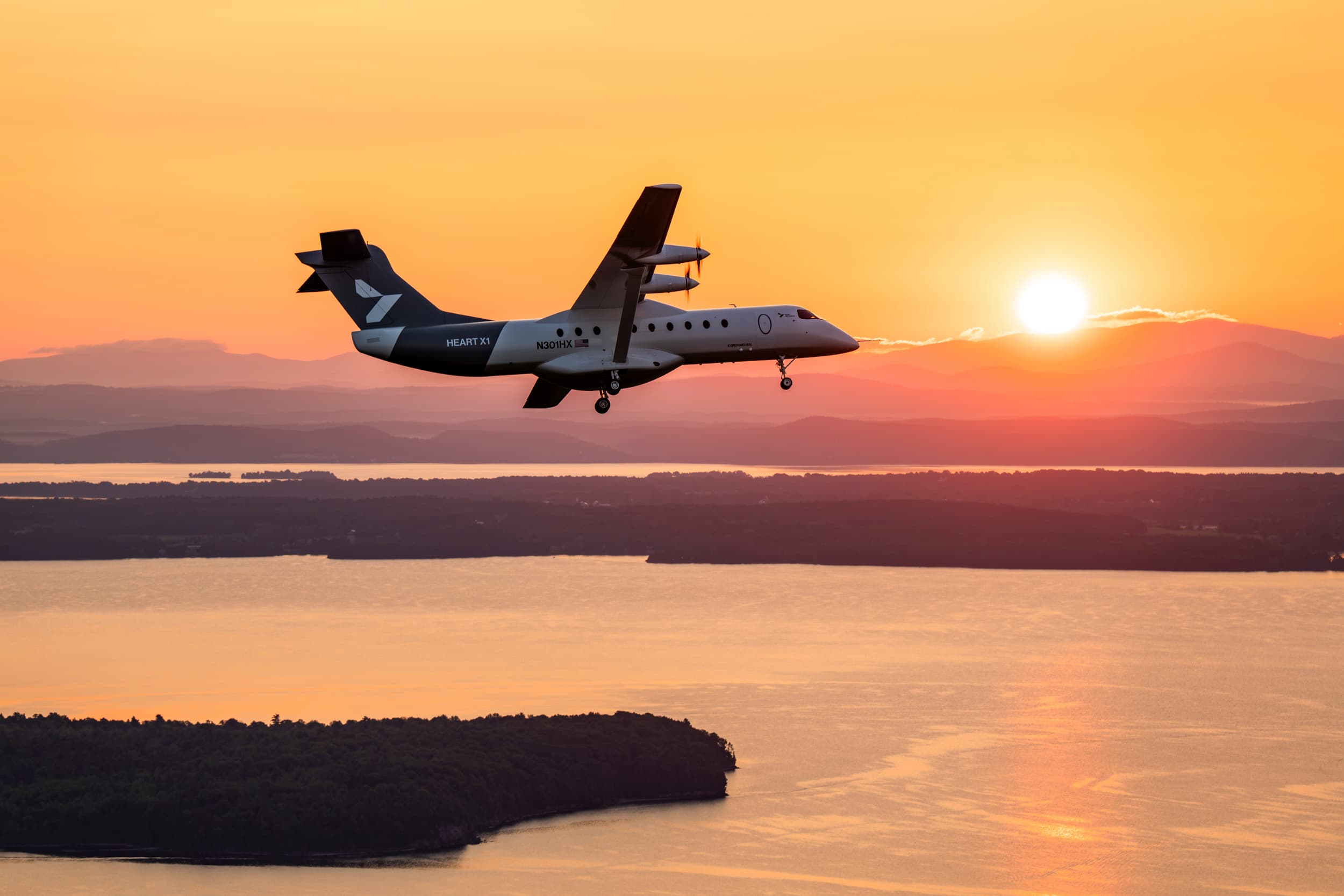 Heart Aerospace’s X1 electric demonstrator flies above Lake Champlain at sunrise with its landing gear extended and Vermont’s Green Mountains beyond.